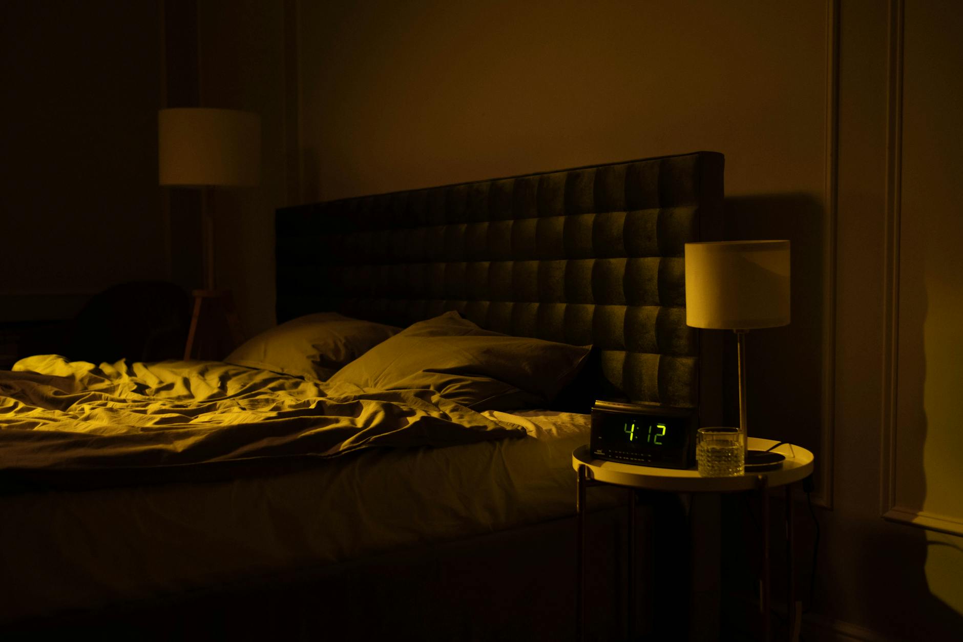 Relaxing dimly lit bedroom with an unmade bed, digital clock displaying 4:12, and warm ambient lighting.