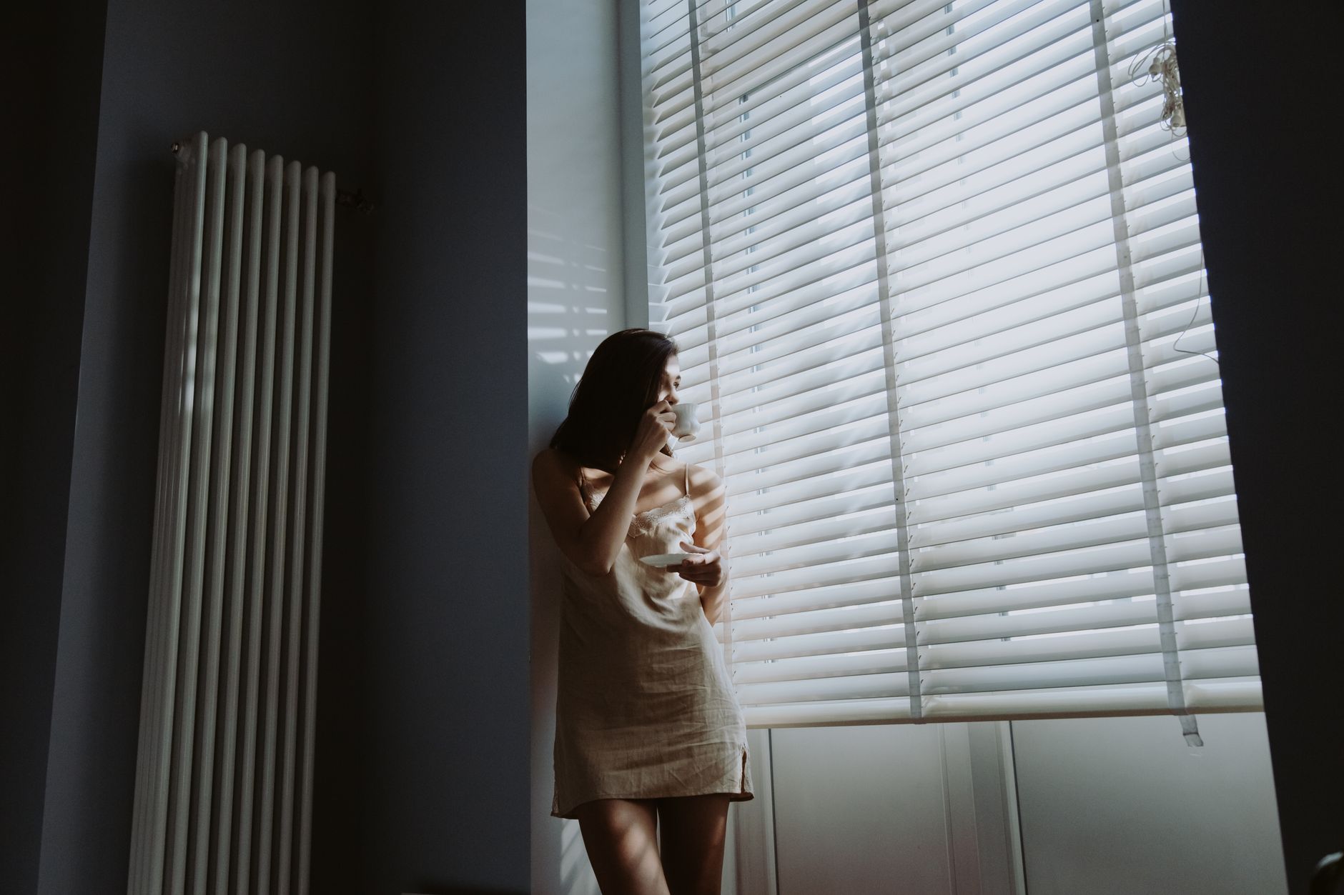 A woman in soft morning light stands by the window, holding a cup and saucer, savoring a peaceful moment.