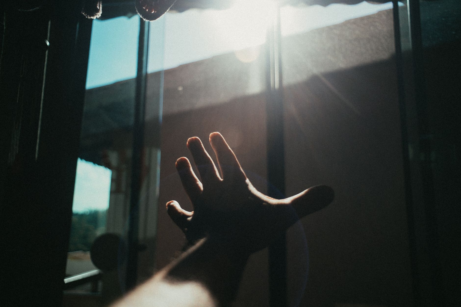 A hand reaches towards the bright sunlight streaming through a window indoors.