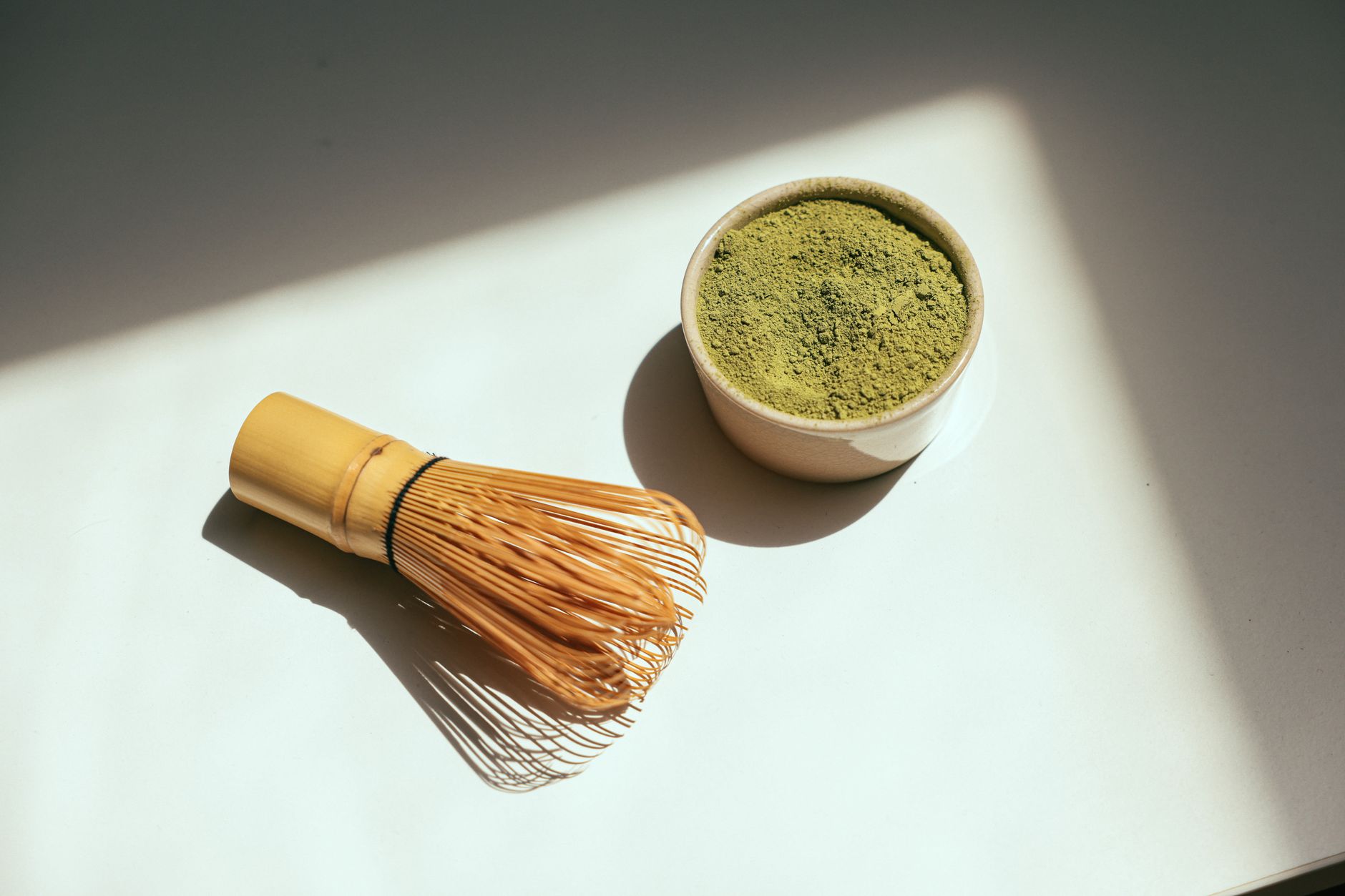 Minimalist composition featuring matcha powder and bamboo whisk in sunlight.