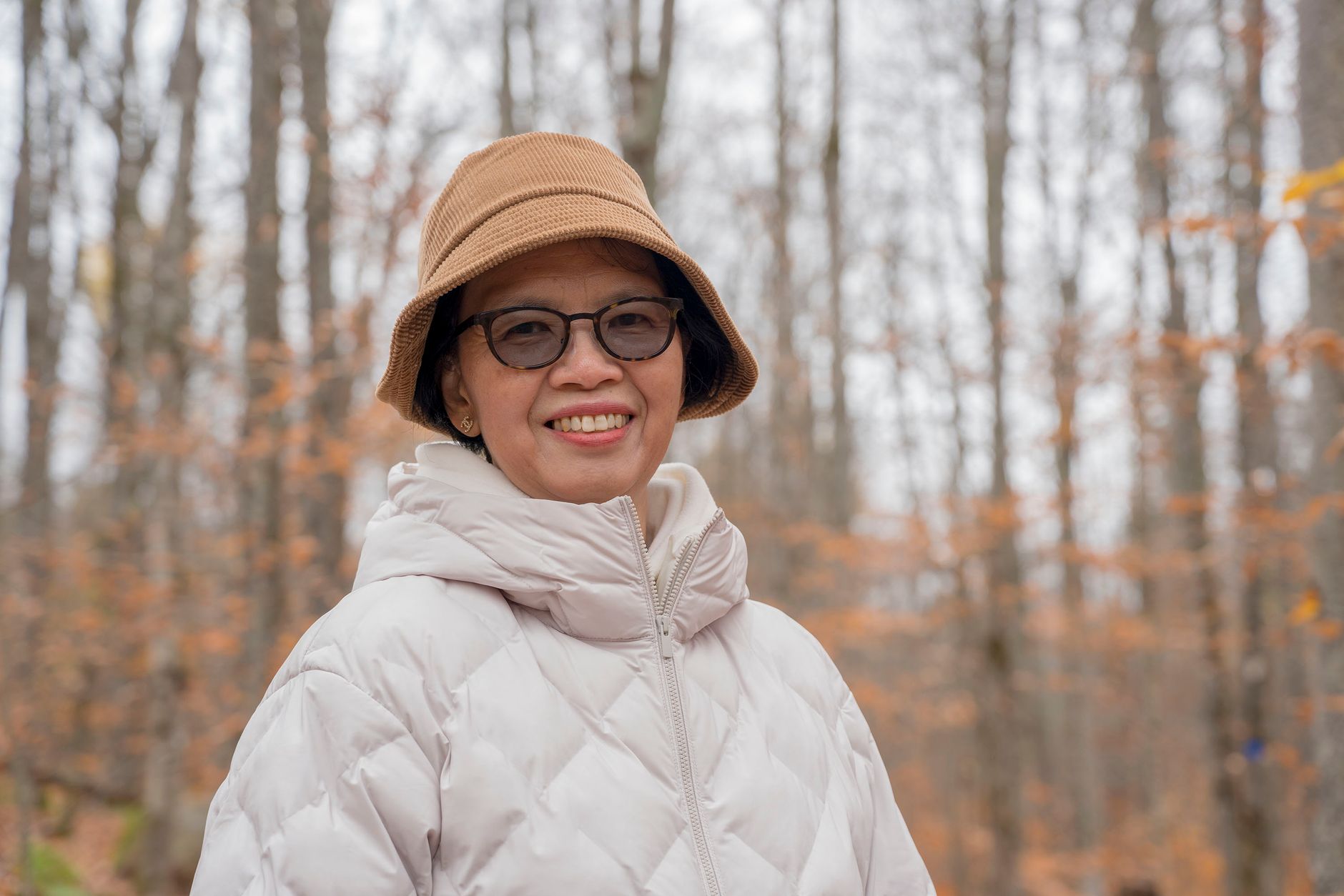 A woman in a hat and jacket smiles warmly in a serene autumn forest setting.